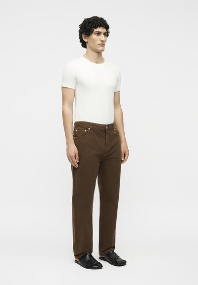 Άνδρας με σγουρά μαλλιά που φοράει εφαρμοστή λευκή κοντομάνικη πουκάμισα, καφέ παντελόνια και μαύρα παπούτσια τύπου slip-on, στέκεται μπροστά σε απλό φόντο.