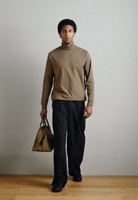 Pruun pikkade varrukatega kõrge kaelusega särk, mustad laiad püksid, hoiab heledat pruuni kandekotti. Minimalistlik kujundus, sile tekstuur.