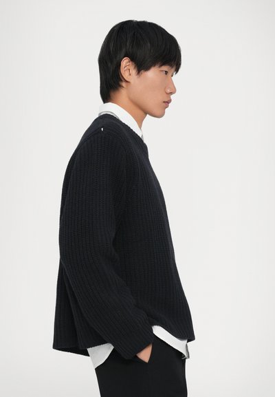 Giovane in maglione nero e camicia bianca, in piedi di profilo con le mani in tasca su uno sfondo bianco uniforme.