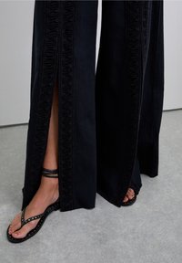 Zwarte wijde broek met gerimpelde zijnaad; heeft uitsparingen voor beweging. Gecombineerd met zwarte platte sandalen met studs.