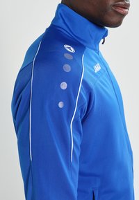 Blauwe sportjacket van gladde stof, met een hoge kraag en witte accenten. Opvallend stippenpatroon op de schouder met merlogo.