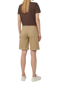 Shorts en coton beige avec des ourlets retroussés, dotés de poches arrière, portés avec un t-shirt marron et des baskets blanches. Modèle présenté de dos.