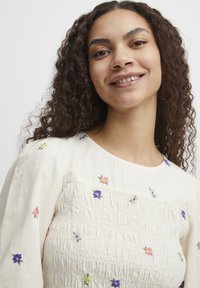 Robes couleur crème avec des motifs floraux brodés en rose, violet et vert. Présente une texture smockée et un col rond avec des manches bouffantes.