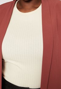 Ljuskrämig ribbad topp med ett scalloped halsband, lagd under en rostfärgad öppna-fram jacka, som visar en slät textur.