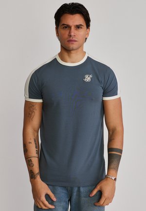 Man with dark hair wearing a blue and white short-sleeve t-shirt and jeans, displaying multiple tattoos on both arms.
