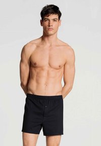 Zwarte katoenen shorts met een elastische tailleband en een enkele knoopsluiting, gedragen door een jonge mannelijke model zonder shirt.