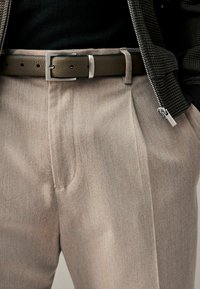 Beżowe eleganckie spodnie uszyte z tkaniny, z płaskim przodem i podwiniętym dołem. Stylizację dopełnia ciemnobrązowy skórzany pasek z srebrną klamrą.