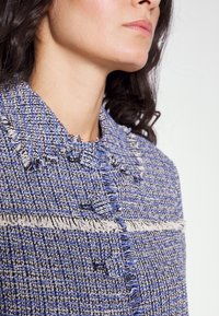 Blå och vit texturerad jacka med fransdetaljer, med krage och två framknappar, som visar ett vävt mönster.