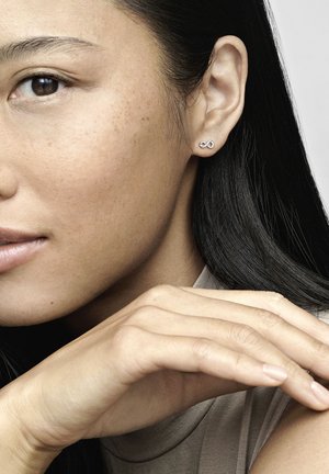 Srebrni uhani v obliki neskončnosti, z gladko površino, ki jih nosi model z srednje dolgimi temnimi lasmi in oblačili v nevtralnih tonih.