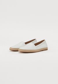 Bela slip-on obuća s ravnim, pletenim jutenim đonom. Gornjište od glatke kože s suptilnim V-oblikovanim izrezom s svake strane za prozračnost.