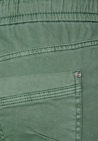 Tejido verde con una textura suave, presenta un bolsillo trasero cosido con hilo a juego y un acento de botón plateado. Diseño minimalista.