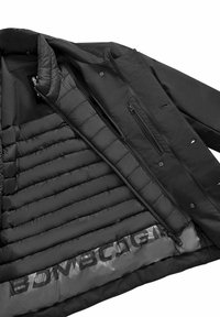 Giacca nera con interno imbottito trapuntato, tasche con zip e una superficie esterna liscia. Logo visibile lungo la fodera interna.