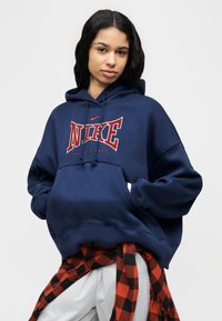 Young woman wearing a navy blue Nike hoodie and a red-black plaid shirt tied around her waist, standing with hands in pockets.
