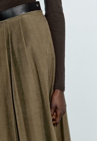 Gonna plissè verde oliva con un finish lucido, dotata di una cintura in pelle nera e dettagli con zip. È visibile una maglietta a maniche lunghe marrone.