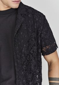 Camicetta in pizzo nero con motivi floreali, maniche corte e un leggero lucido, indossata sopra una maglietta di cotone scura.