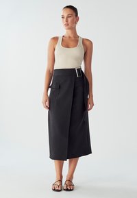 Žebrovaný béžový top na ramínka v kombinaci s černou midi sukní s opaskem, postranními kapsami a elegantním, upraveným střihem. Jednoduché černé sandály.