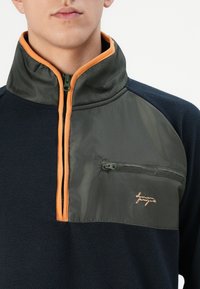 Pullover in felpa con colletto alto, chiusura a mezza zip e tessuto blu navy. Presenta un pannello superiore verde e bordature arancioni, con una tasca sul petto con zip.
