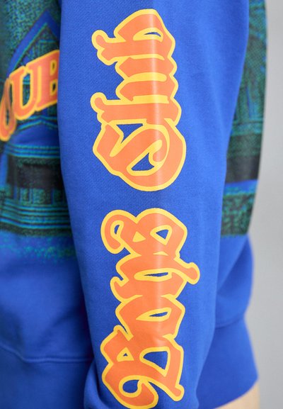 Blå hettegenser med kraftig oransje tekst som leser "Club" og "Bang" i stilisert skrift på ermene, med et fargerikt mønsterdesign.