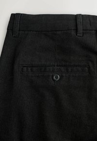 Schwarze Denim-Hose mit einer traditionellen Gesäßtasche, einem einzelnen schwarzen Knopf und genähten Details entlang der Kanten.