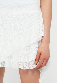 Το χέρι μιας γυναίκας κρατάει απαλά το τελείωμα μιας λευκής δαντελένιας φούστας με λουλουδάτα σχέδια και βολάν στο τελείωμα.