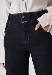 Donkerblauwe denim high-waisted jeans met een sluiting met drie knopen, oranje stiksels en zijzakken. Gepaard met een wit overhemd met lange mouwen.