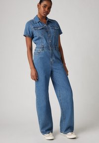 Denim jumpsuit med korte ermer, kragehals, frontknapper, to brystlommer og vid benpassform i mellomblå vask.