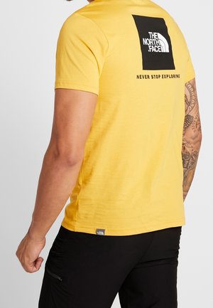 Mand iført gul The North Face t-shirt med sort logo og teksten "Never Stop Exploring" på ryggen, sorte bukser og synlige tatoveringer på armene.