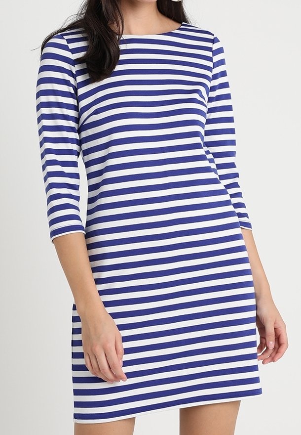 Femme portant une robe mi-longue à rayures horizontales bleues et blanches avec des manches trois-quarts, debout sur un fond uni.