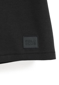 Zwarte katoenen T-shirt met een getextureerd zwart label waarop "POLO CLUB" is geperst. De zoom heeft een schone afwerking met zichtbare stiknaden.