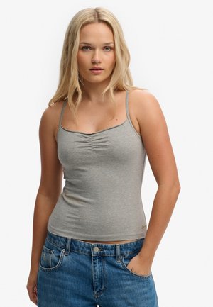 Młoda kobieta z długimi blond włosami, ubrana w szarą marszczoną kamizelkę i niebieskie dżinsy, stojąca z jedną ręką w kieszeni.