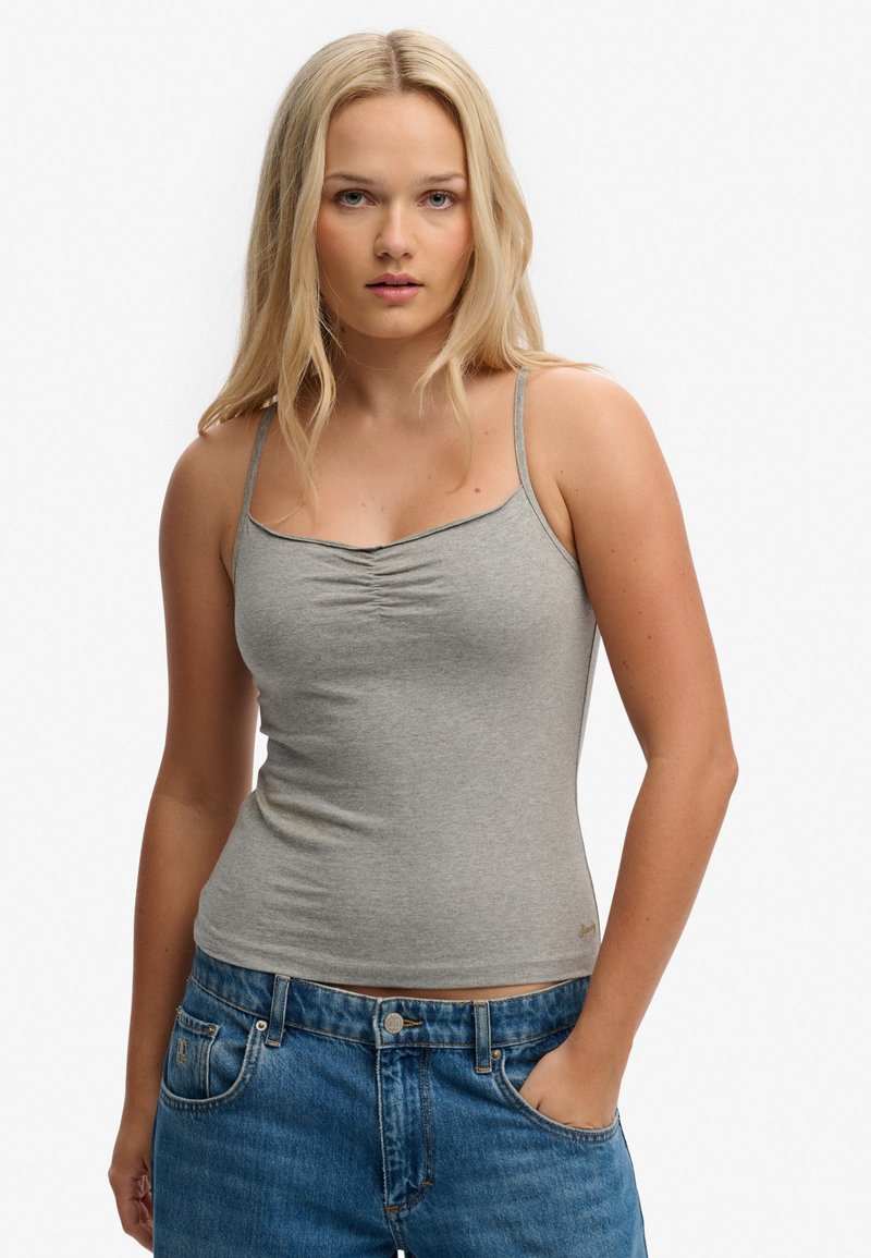 Joven mujer con cabello largo y rubio, vestida con un camisole gris fruncido y jeans azules, de pie con una mano en el bolsillo.