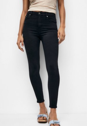 Femme portant un jean skinny noir taille haute et des sandales ouvertes à talons bleu clair, debout devant un fond blanc.