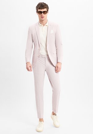 Helles rosa Anzug mit einem einreihigen Sakko, schmal geschnittenen Hosen und einem cremefarbenen Poloshirt. Getragen mit beigen Sneakers und quadratischen Sonnenbrillen.
