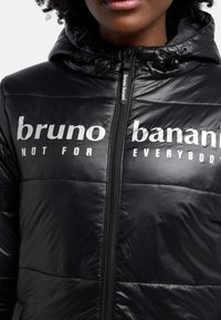 Svart vadderad jacka med glänsande textur, med dragkedja och huva. Tryckt text läser "bruno banani NOT FOR EVERYBODY" i vitt.