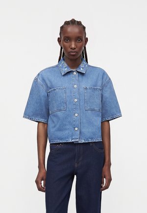 Žena s vrkôčikmi má na sebe svetlomodrú rifľovú košeľu s krátkym rukávom, vyšívaným kvetinovým golierom a tmavomodré rifle.