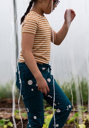 Young girl with braided hair wearing glasses, striped orange shirt, and blue pants walks among hanging white strings in a garden setting.