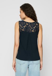 Top sans manches bleu marine avec un dos en dentelle florale, affichant une coupe décontractée et un détail plissé à l'arrière. Le tissu semble doux et léger.