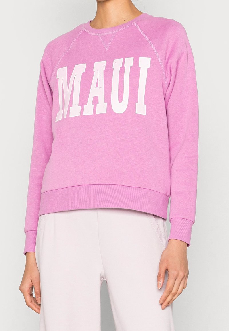 Personne portant un sweat-shirt rose avec « MAUI » en grandes lettres blanches et un pantalon clair, cadré au-dessus du menton et à mi-cuisse.