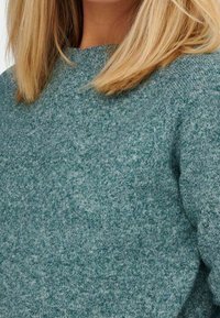 Türkisfarbener Strickpullover mit einer weichen, texturierten Optik. Mit Rundhalsausschnitt und langen Ärmeln. Der Stoff hat ein leicht geflecktes Muster.