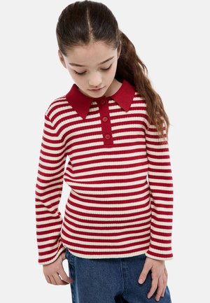 Fille aux longs cheveux portant un pull rayé rouge et blanc avec un col rouge et des boutons, regardant vers le bas avec les mains près des hanches.