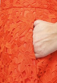 Orange spetsklänning med blommig broderi, med en synlig ficka som är vackert inramad med intrikata sömmar och en mjuk, något genomskinlig textur.