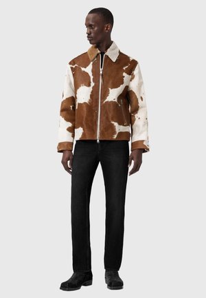 Mees, kes kannab pruunide ja valgete mustritega lukuga jope, musti teksapükse ja musti saapaid, seistes ühevärvilise helehalli tausta ees.