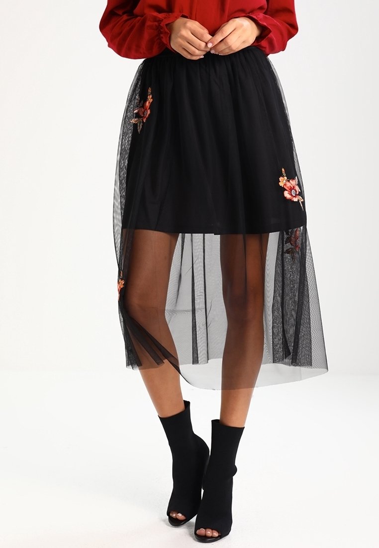 Femme portant une jupe midi noire en dentelle florale transparente avec une sous-jupe noire unie et des bottines noires ouvertes à l'avant.