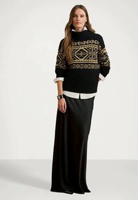 Kvinne i en svart og gullmønstret genser over en hvit skjorte, kombinert med et langt svart skjørt, som står foran en ensfarget bakgrunn.