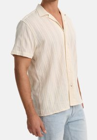 Homme portant une chemise crème texturée à manches courtes avec col campagnard et des jeans bleu clair, sur fond blanc.