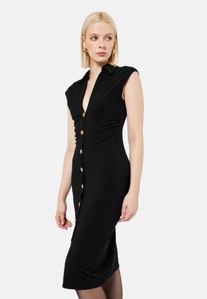 Kobieta o blond włosach, ubrana w czarną, bezrękawnikową sukienkę midi z guzikami i kołnierzykiem, stojąca na jednolitym jasnym tle.