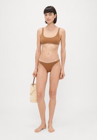 Smeđi bikini od rastezljivog materijala s kontrastnim bijelim šavovima. Model drži pleteni slamnati koš, stojeći bos protiv jednostavnog pozadina.