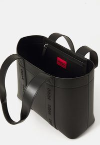 Sac fourre-tout en cuir noir avec deux anses larges, logos HUGO en relief sur les sangles avant, et une poche zippée intérieure avec une étiquette HUGO rouge.