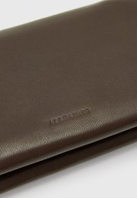 Portafoglio in pelle marrone con texture liscia, logo impresso e design sottile. Presenta linee pulite e un'estetica minimalista.