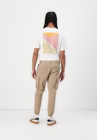 Man met gevlochten haar, gekleed in een wit T-shirt met opdruk, beige cargobroek met elastische boorden, witte sokken en witte sneakers met zwart-witte strepen, gekeerd van de camera.